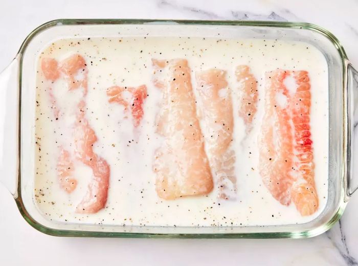 A glass dish containing catfish fillets submerged in a seasoned buttermilk mixture.