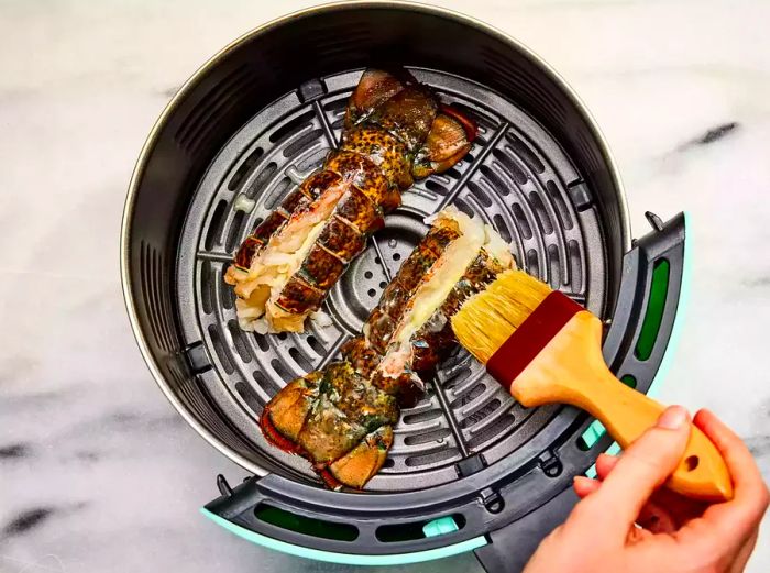 An air fryer basket with two lobster tails being coated with garlic-lemon butter