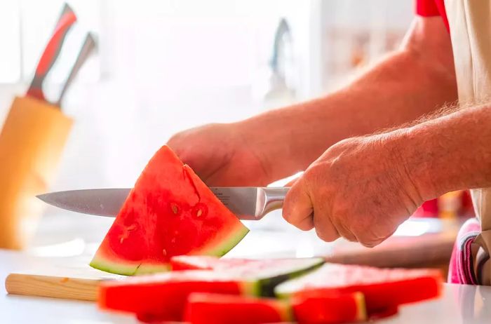 An elderly man cutting a ripe red watermelon in a home kitchen.