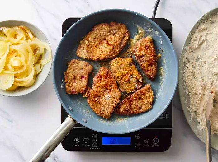 A large skillet with flour-coated liver slices frying in butter