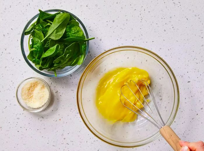 A glass bowl with eggs being whisked by a metal whisk, accompanied by a bowl of fresh baby spinach and a dish of Parmesan cheese