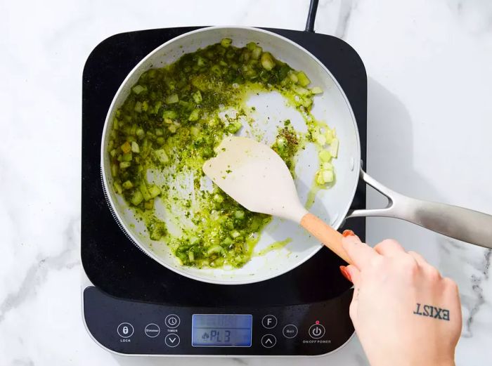 A skillet with sautéed onions, pesto, salt, and pepper being stirred with a spatula