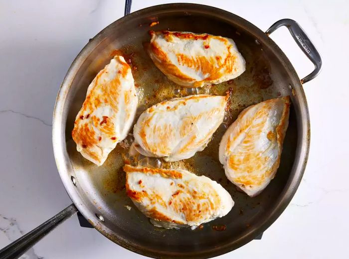 Five chicken breasts frying in a skillet.