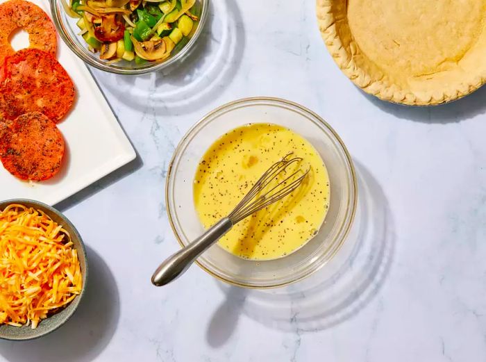 A bowl of whisked eggs, milk, salt, and pepper, accompanied by cooked vegetables, fried tomato slices, shredded cheese, and a pie crust