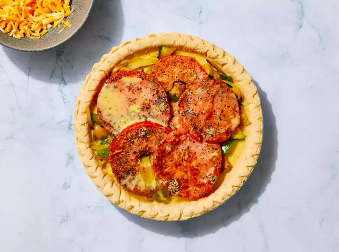 A pie crust filled with layers of shredded cheese, whisked eggs, sautéed vegetables, and topped with golden-brown tomato slices