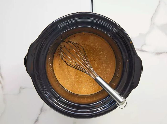 A slow cooker insert with beef broth, condensed soup, steak sauce, Worcestershire sauce, garlic powder, paprika, salt, and pepper being mixed together with a whisk.