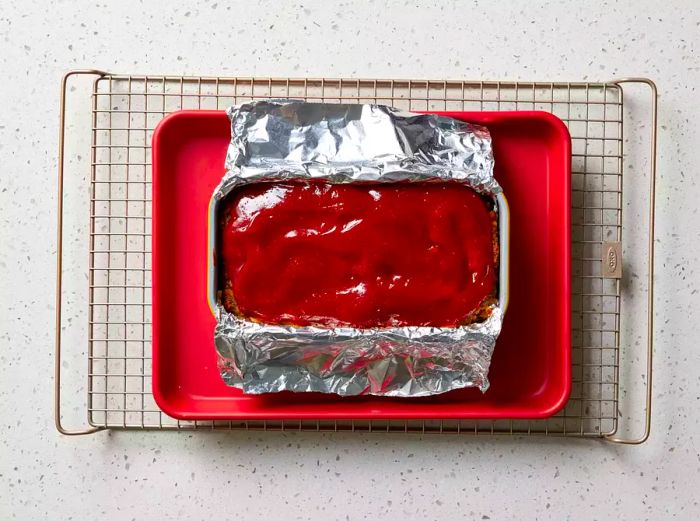 Stovetop Stuffing Meatloaf resting in a loaf pan with aluminum foil, placed on a baking sheet and cooling rack