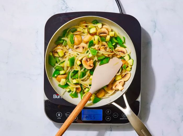 A skillet filled with sautéing onion, bell pepper, mushrooms, and zucchini, being stirred with a rubber spatula