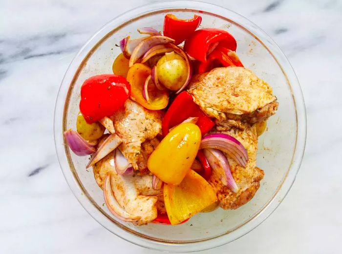 Chicken thighs, onions, bell peppers, and potatoes are added to the bowl and stirred to coat evenly.