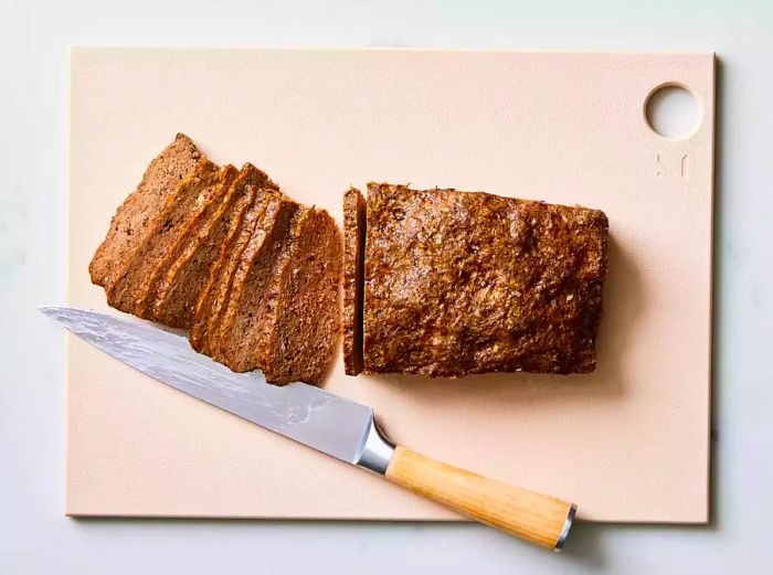 A loaf of homemade gyro meat placed on a cutting board, with thin slices next to a knife