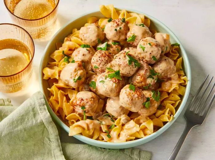 A bowl of slow-cooked Swedish meatballs served on top of noodles, garnished with chopped parsley.