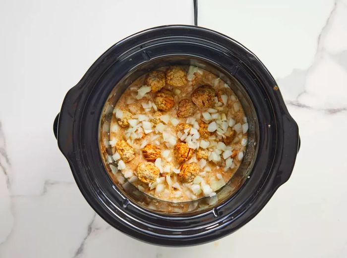 A slow cooker insert with meatballs, diced onions, and brown gravy.