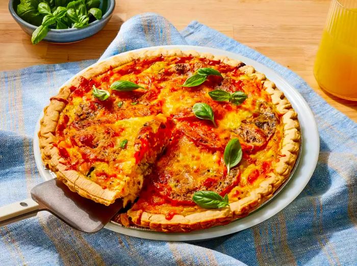 A loaded vegetarian quiche garnished with fresh basil leaves, with a slice being carefully removed