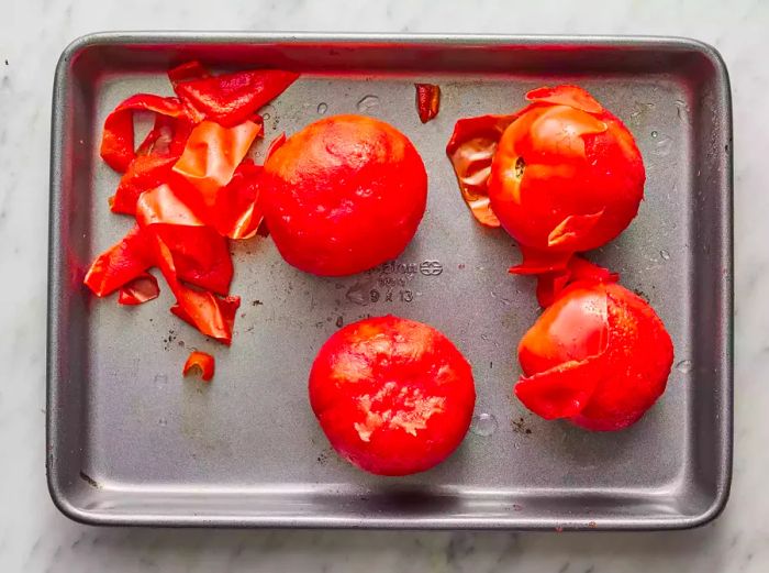 Blanched tomatoes laid out on a baking sheet, with the skin being peeled off.