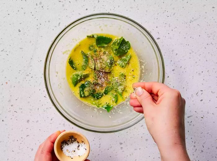 A hand sprinkling salt into a bowl of whisked eggs, mixed with baby spinach and Parmesan cheese