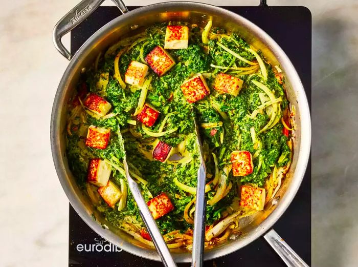 A skillet of saag paneer being stirred with tongs