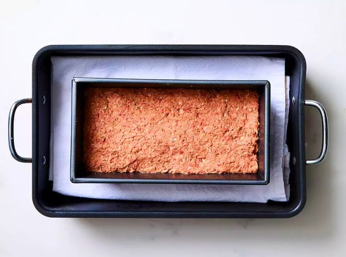 A loaf pan filled with the processed meat mixture, placed in a towel-lined baking pan