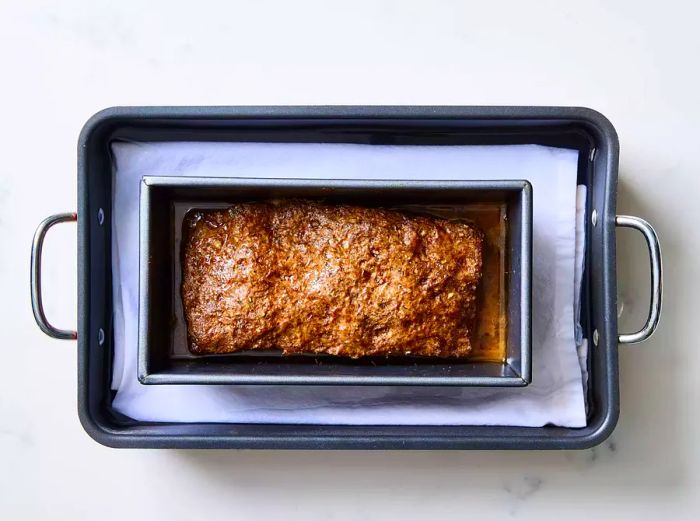 A loaf pan containing baked gyro meat, resting in a roasting pan lined with a towel and filled with water