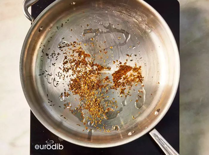 A large skillet with cumin seeds sizzling in oil