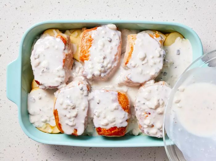 A baking dish with layers of sliced potatoes, topped with seared pork chops and onions, all covered with the soup and milk mixture.