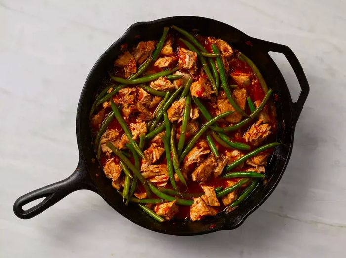 Aerial view of green beans being mixed and cooked with pieces of pork and sauce in a skillet.