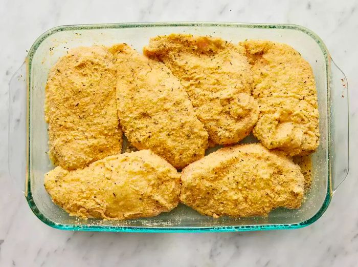 A top-down view of breaded chicken breasts resting in a glass baking dish, ready to be baked.