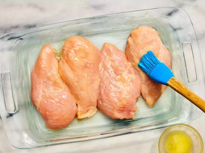 A baking dish with four chicken breasts being brushed with a melted butter and salt blend.