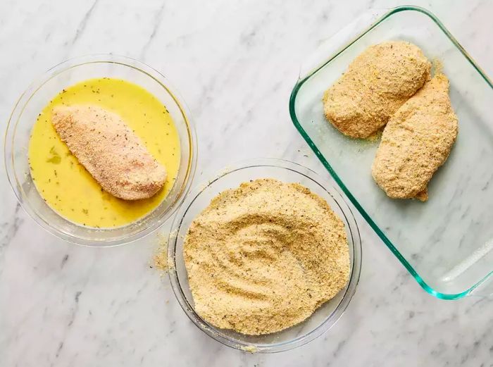 Raw chicken breasts being dipped in an egg wash, coated in breadcrumbs, and arranged in a baking dish.