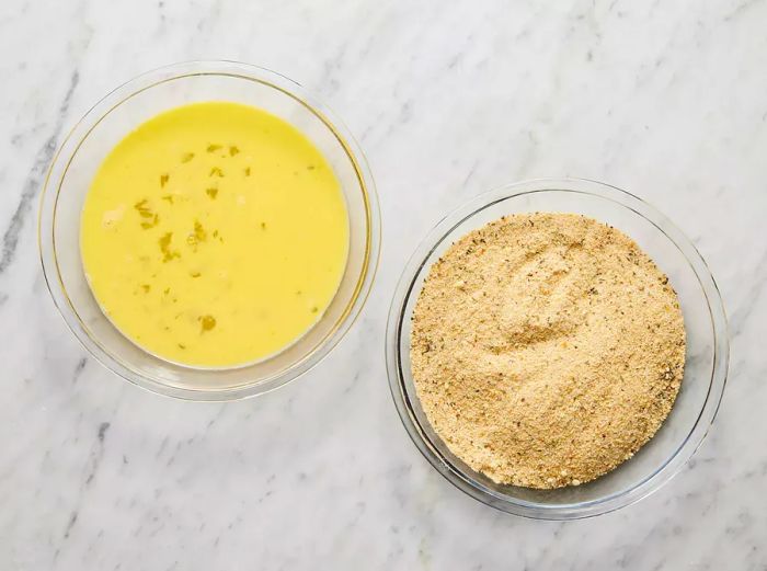 A top-down view of one bowl containing whisked eggs and another with seasoned breadcrumbs.