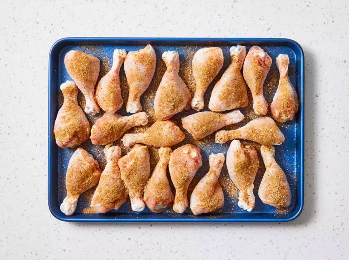 A baking sheet filled with chicken drumsticks seasoned with Cajun spice, poultry seasoning, garlic powder, salt, and black pepper.