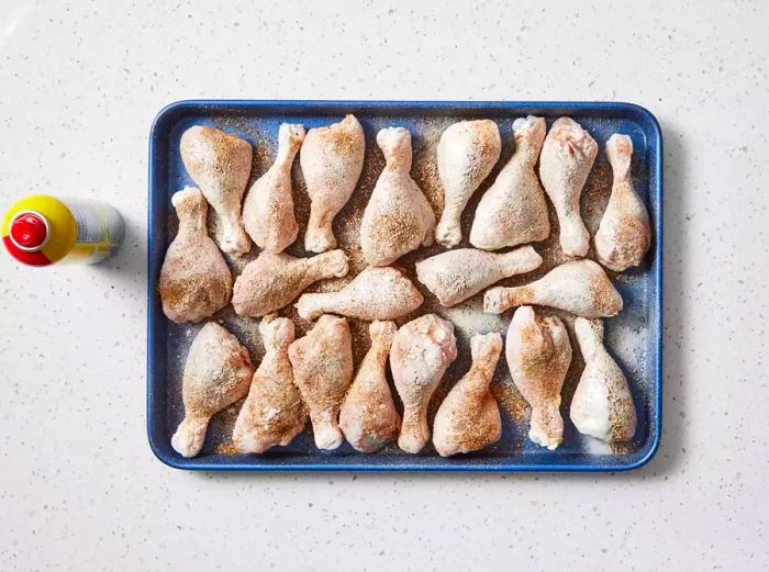 A baking sheet featuring seasoned chicken drumsticks, dusted with flour and coated with cooking spray.