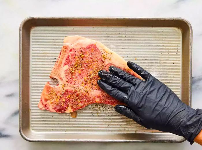 A top-down view of a raw porterhouse steak being seasoned before cooking.