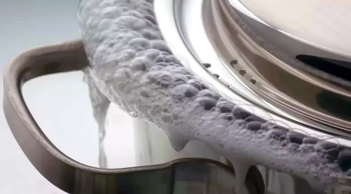 Water boiling over from a saucepan with a lid, creating a mess. Close-up of the pot spilling over.