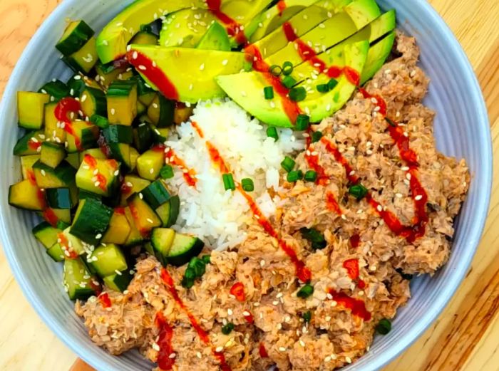 a white bowl filled with rice, canned salmon, cucumber slices, and avocado, drizzled with sriracha sauce