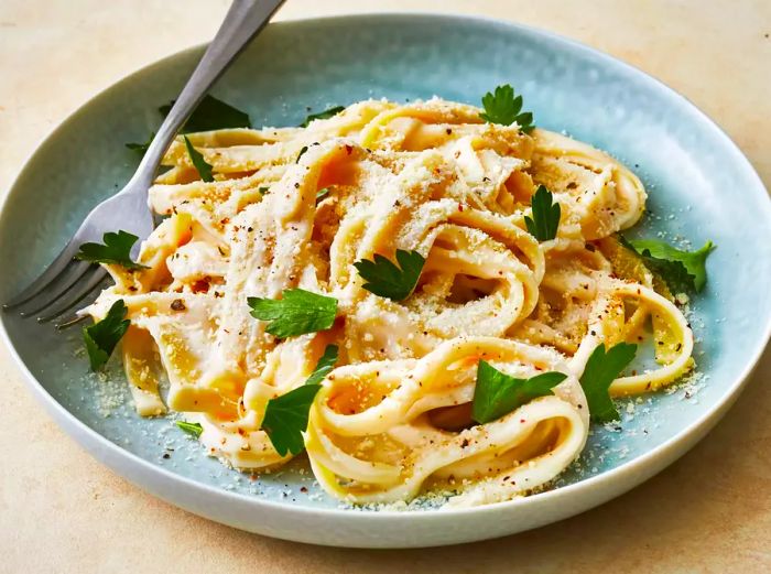 a low-angle, close-up view of a plate of fettuccine pasta smothered in creamy Alfredo sauce, topped with fresh parsley and a sprinkle of black pepper