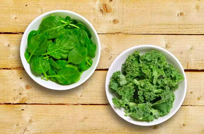 Two white bowls, one filled with spinach leaves and the other with chopped kale, shot from above on a rustic wooden surface