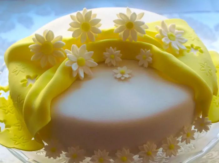 A cake adorned with a flawless white fondant covering, accented with a yellow fondant ribbon and delicate daisy decorations.