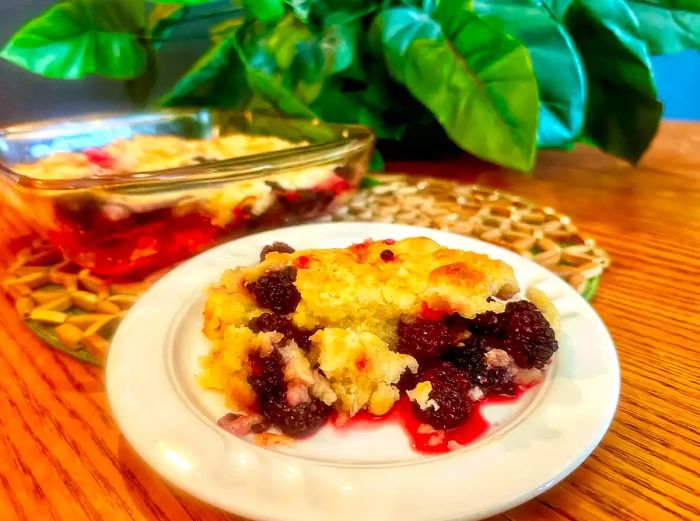 A serving of blackberry cobbler on a white plate, with the baking dish in the background