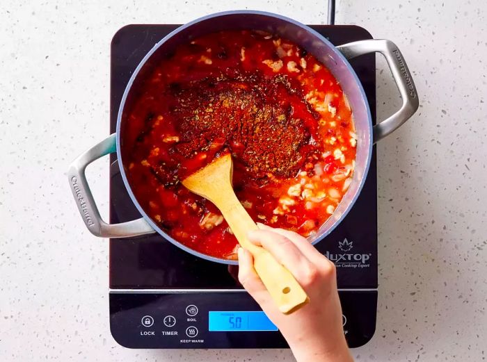 A wooden spoon stirring chili powder, paprika, oregano, cayenne pepper, cumin, salt, and black pepper into a pot of simmering chili.