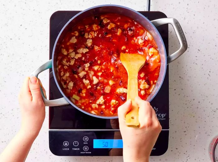 A large pot with beans, crushed tomatoes, and garlic being stirred together with the browned ground turkey.