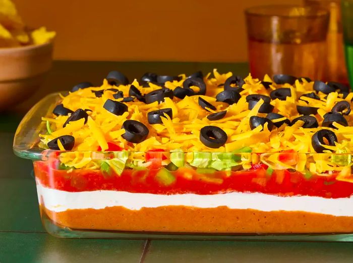 A glass baking dish filled with seven-layer taco dip, topped with shredded cheese and sliced black olives.