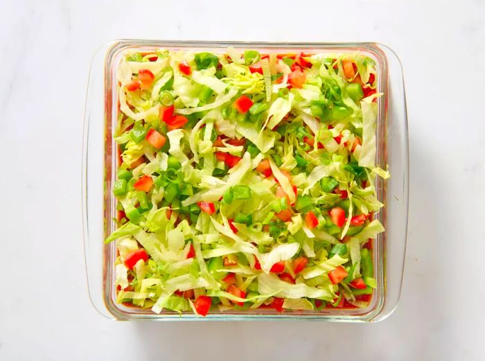 A glass baking dish layered with refried beans, sour cream-cream cheese mixture, salsa, lettuce, tomatoes, and bell peppers.