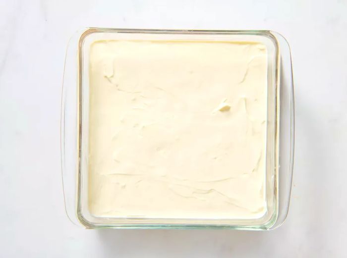 A glass baking dish with a layer of sour cream and cream cheese spread over the refried beans.