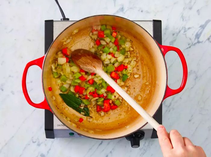 Overhead shot of hand stirring bay leaves and spices into the mirepoix with a wooden spoon