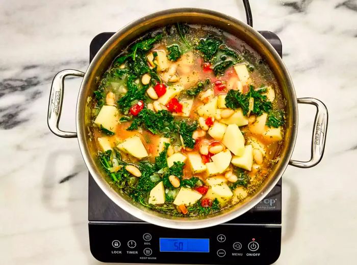 A large pot of soup brimming with beans, potatoes, kale, and aromatic seasonings