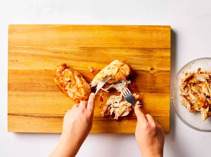 A cutting board with chicken breasts being shredded with two forks.