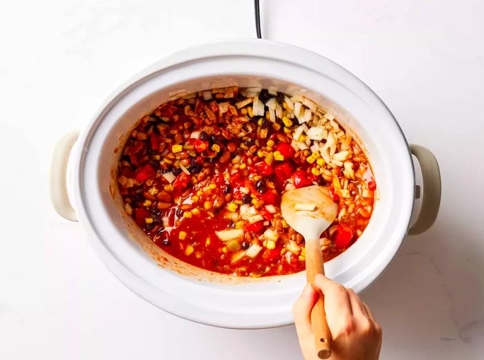 A slow cooker filled with diced tomatoes, chili beans, black beans, corn, beer, tomato sauce, and onion, all being stirred together with a spatula.