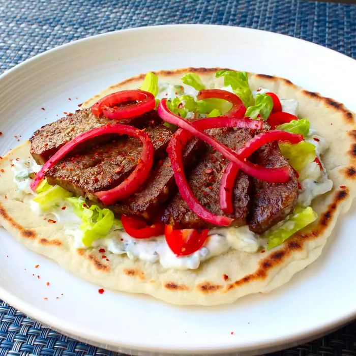 A close-up shot of an American Gyro served on a pita, topped with red onions, lettuce, and tomatoes, on a plate.