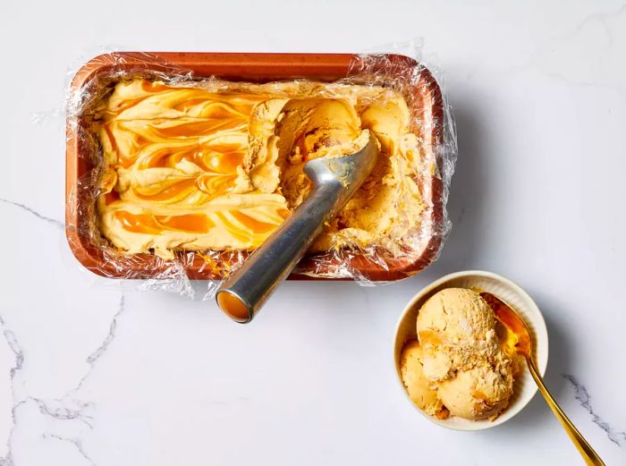 Scoops of Dulce De Leche Ice Cream topped with flaky sea salt, served in a bowl with a spoon, alongside a loaf pan of ice cream and a scoop