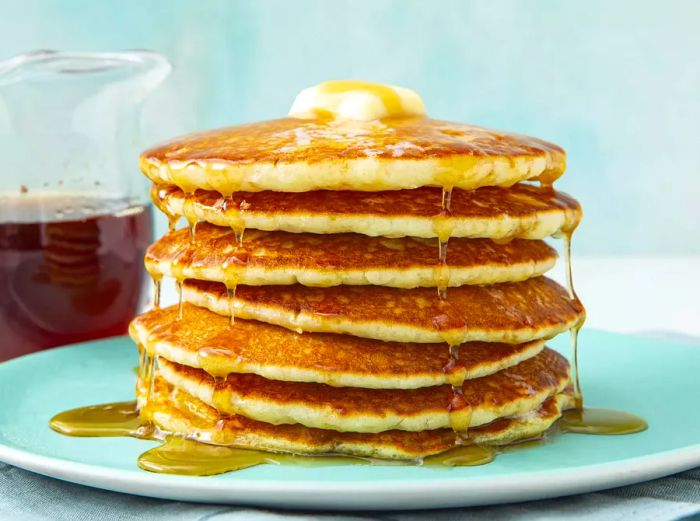 A low-angle view of a stack of golden buttermilk pancakes topped with melting butter and drizzled with syrup